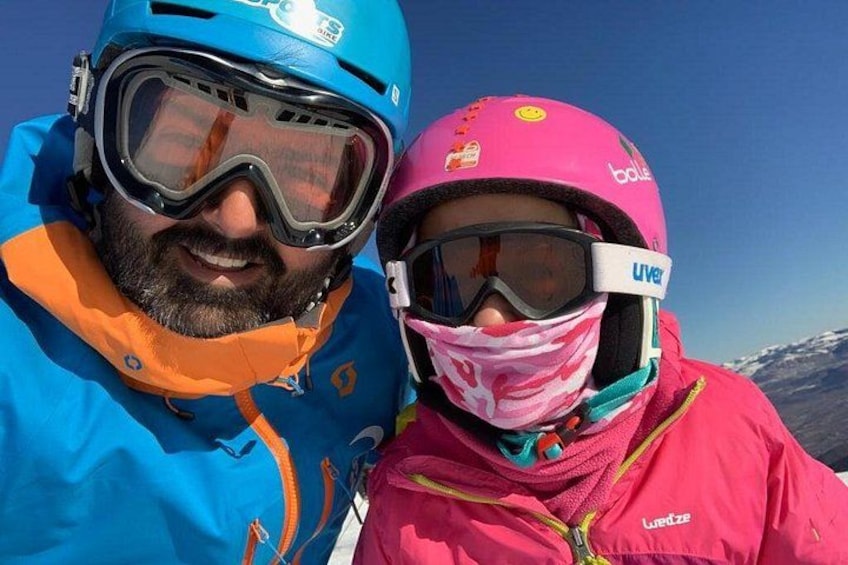 Two people wearing helmets and goggles, one in blue and the other in pink, with a mountainous landscape in the background. 1