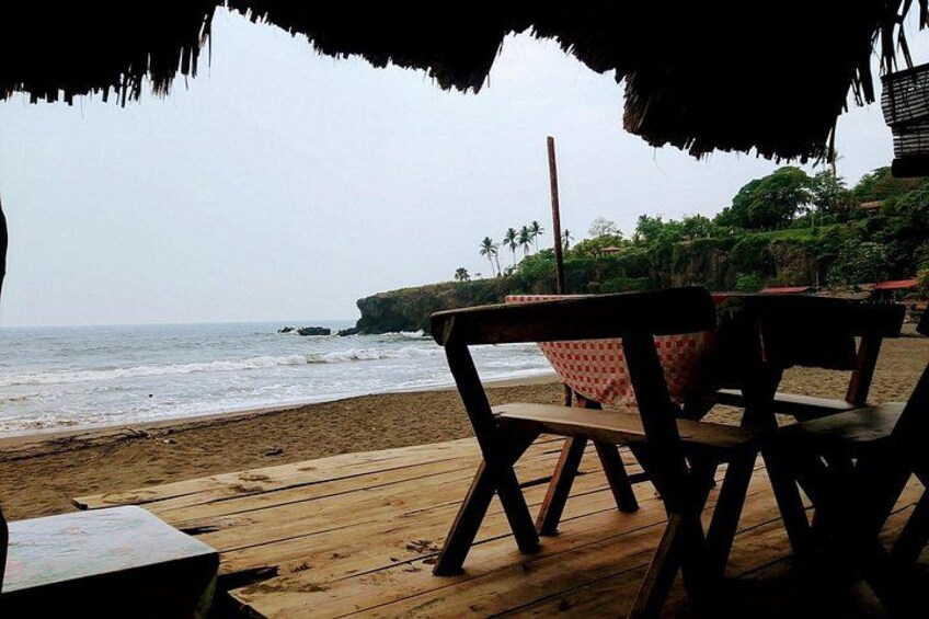 A wooden deck with chairs and a table, overlooking a beach with a thatched roof structure. 4