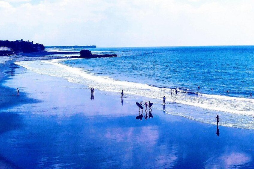 A beach with people walking and standing, a large rock formation, and calm blue waters. 2