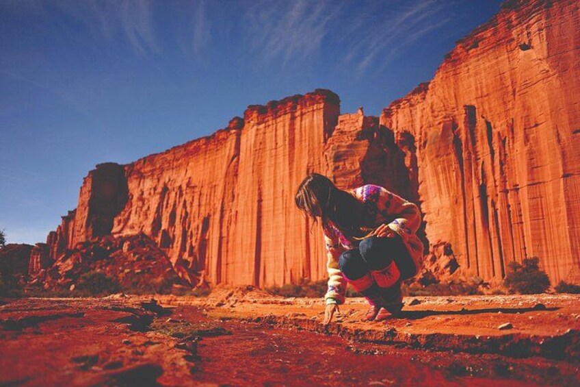 A person in a colorful jacket and hat, bending over in a desert landscape with red rock formations. 1