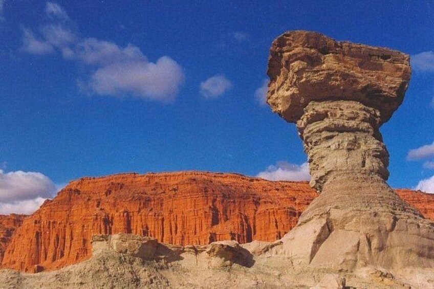 A rock formation with a balanced rock on top, a red cliff in the background, and a clear blue sky. 5