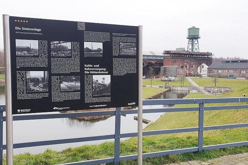 A historical information board by a water body with industrial structures in the background. 4
