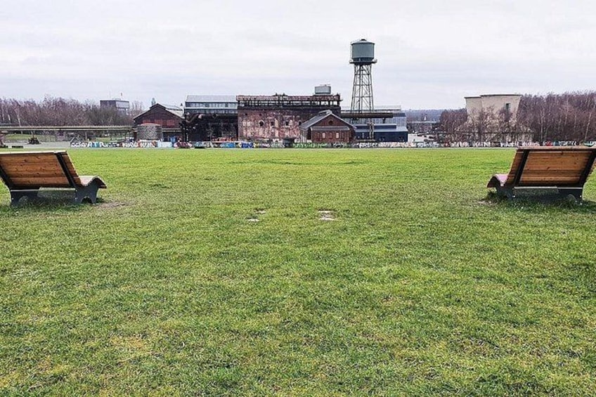 A grassy field with two wooden benches, a water tower, and industrial buildings in the background. 5