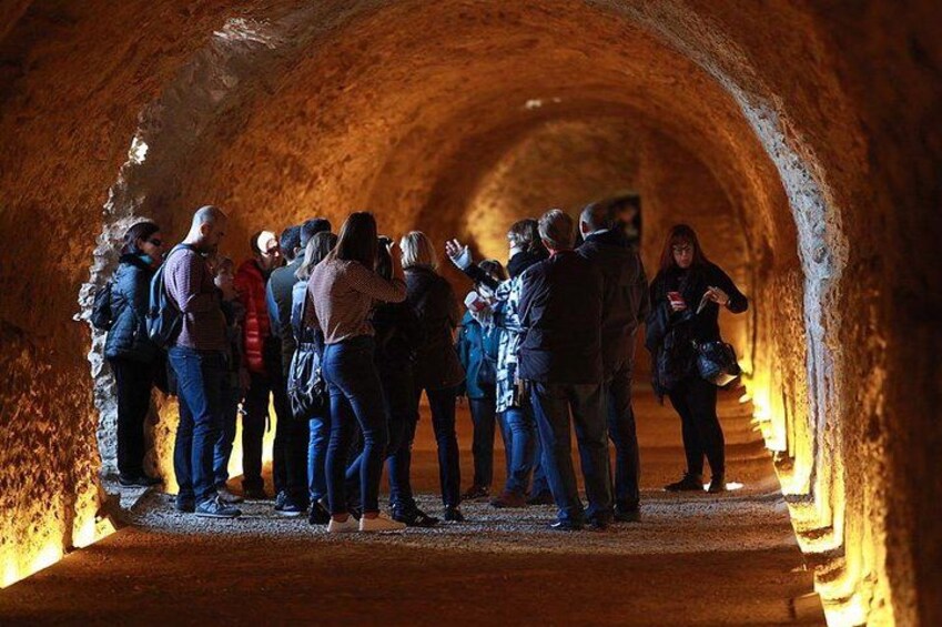 Eine Gruppe von Menschen steht in einem steinernen Tunnel, am Ende ist eine warme Lichtquelle zu sehen. 4