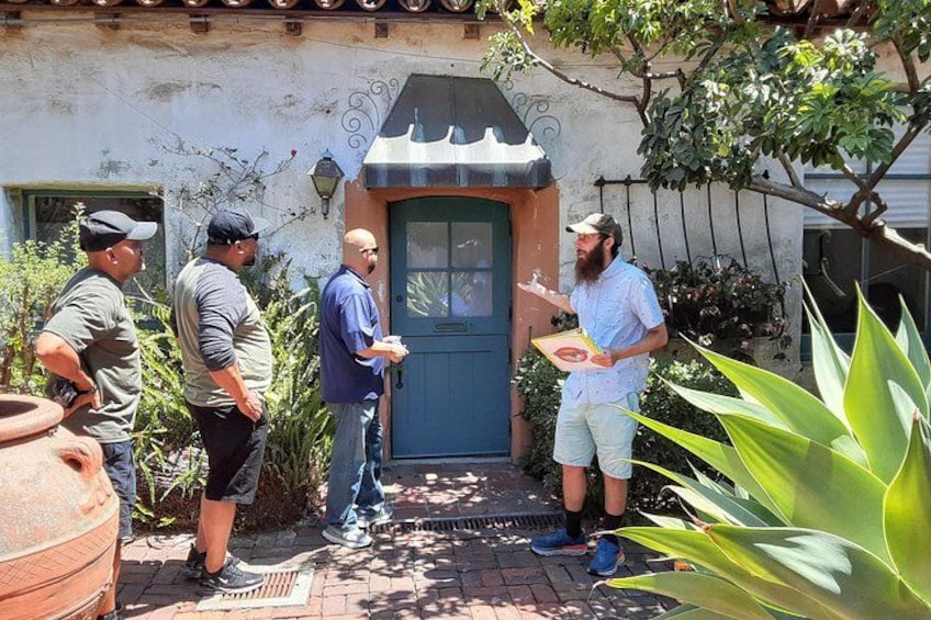Un groupe de personnes se tient devant un bâtiment dont la porte est bleue, entouré de plantes et d’un chemin pavé en briques. 4