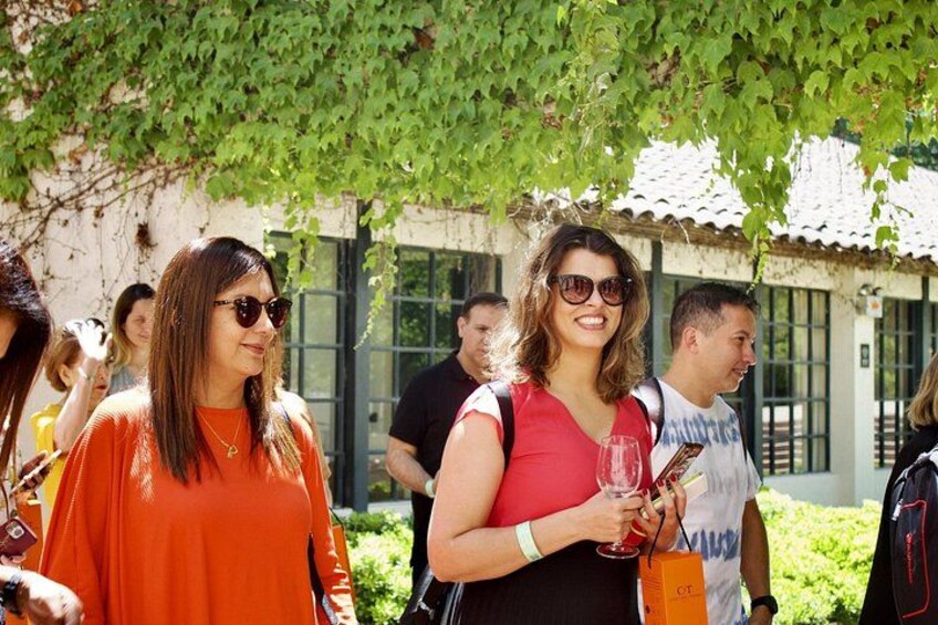 Personas de pie al aire libre, algunas sosteniendo copas de vino, con follaje y un edificio al fondo. 3