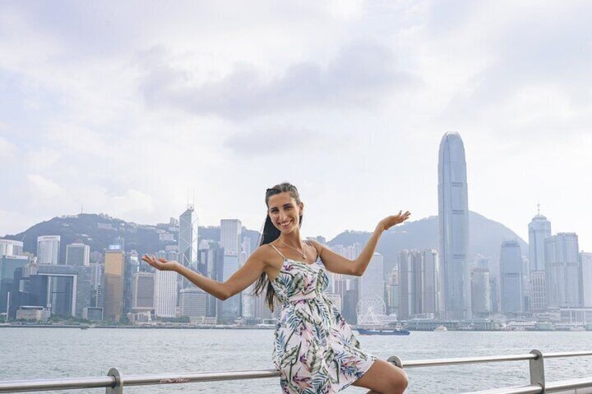 A person in a floral dress posing with arms outstretched in front of a city skyline with prominent skyscrapers and a body of water. 3