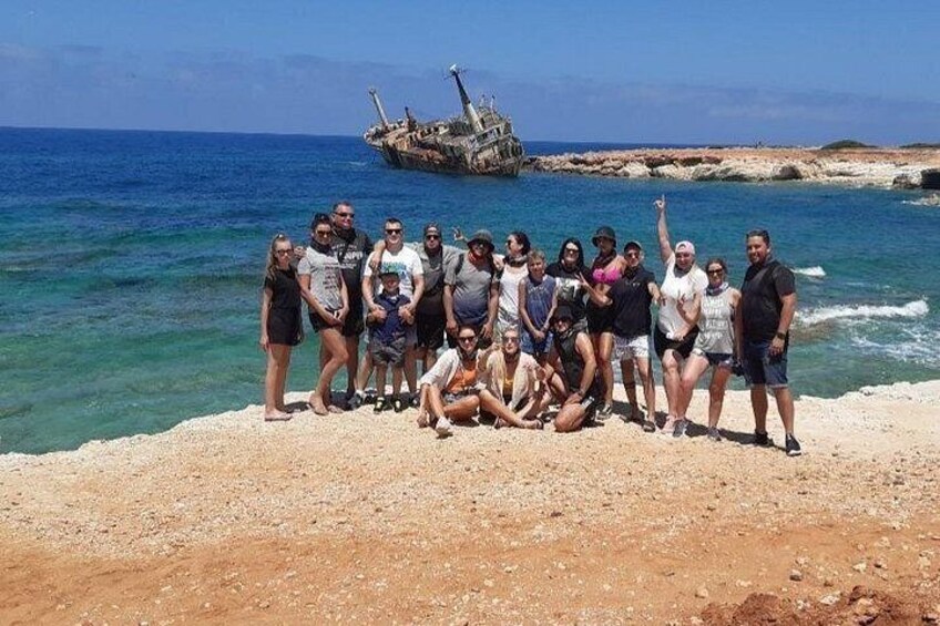 Eine Gruppe von Personen posiert an einem felsigen Strand, im Hintergrund ein Schiffswrack. 1