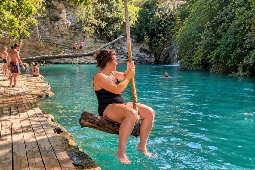 Eine Person in einem schwarzen Badeanzug sitzt auf einer Holzschaukel über türkisfarbenem Wasser, umgeben von viel Grün und Felsen. 2