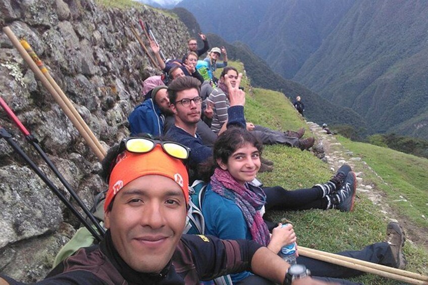 Eine Gruppe von Menschen sitzt auf einem Bergpfad, an einer Seite eine Felswand und im Hintergrund ein Tal. 1