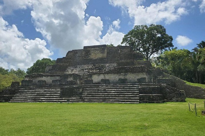 A large ancient pyramid with multiple levels and a staircase leading up to it, surrounded by a grassy area and trees. 1