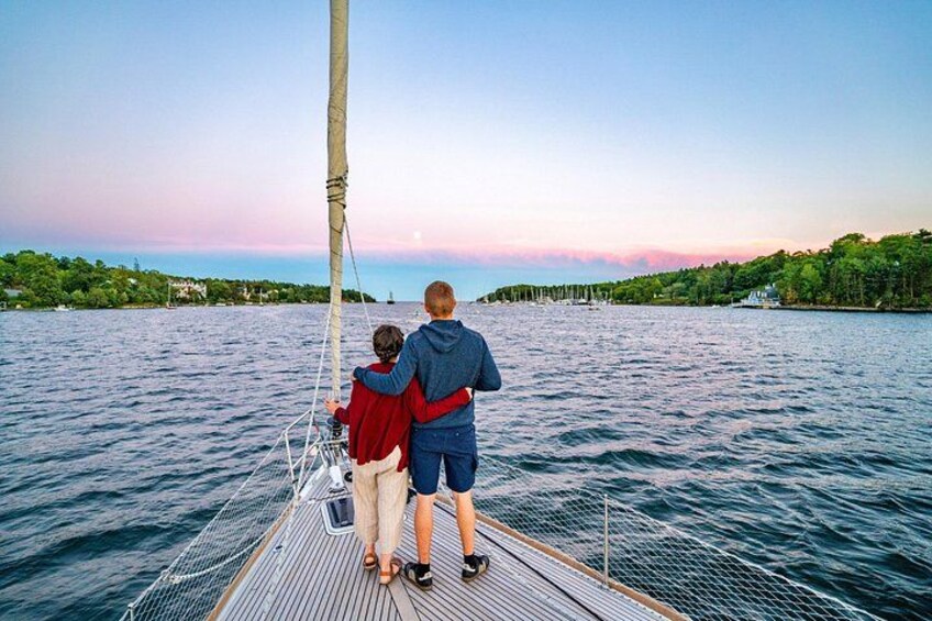 A sailboat on calm water with a person and a child on deck, a clear sky, and a distant shoreline with greenery. 4