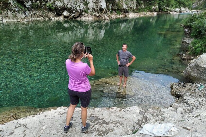A person taking a photo of another person standing in a clear water body surrounded by rocky terrain. 2