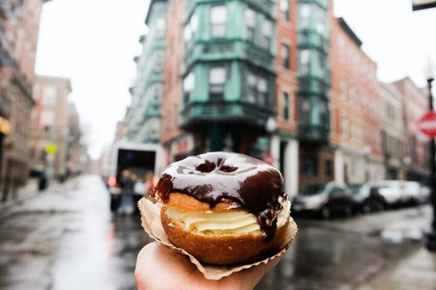 Une main tenant un beignet enrobé de chocolat et fourré à la crème, sur fond d’une rue de la ville avec des bâtiments et un trottoir mouillé. 1