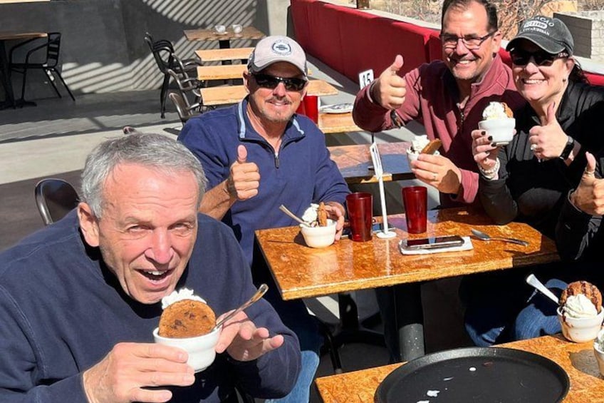 Cuatro personas sentadas en una mesa al aire libre, comiendo postres y haciendo gestos de aprobación. 2