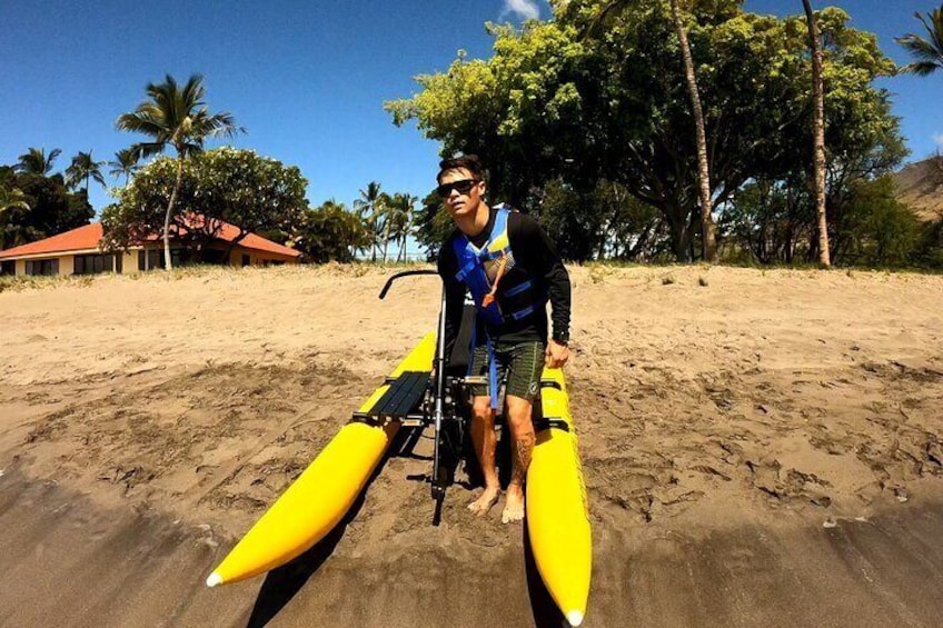 A person standing on a yellow surfboard on a sandy beach with palm trees and a building in the background. 2