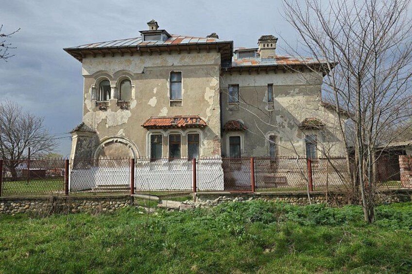 A large, weathered building with arched windows and a metal roof, surrounded by a chain-link fence and overgrown grass. 3