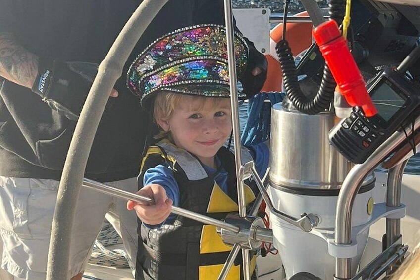 A child on a boat steering wheel, wearing a life jacket and a hat. 3
