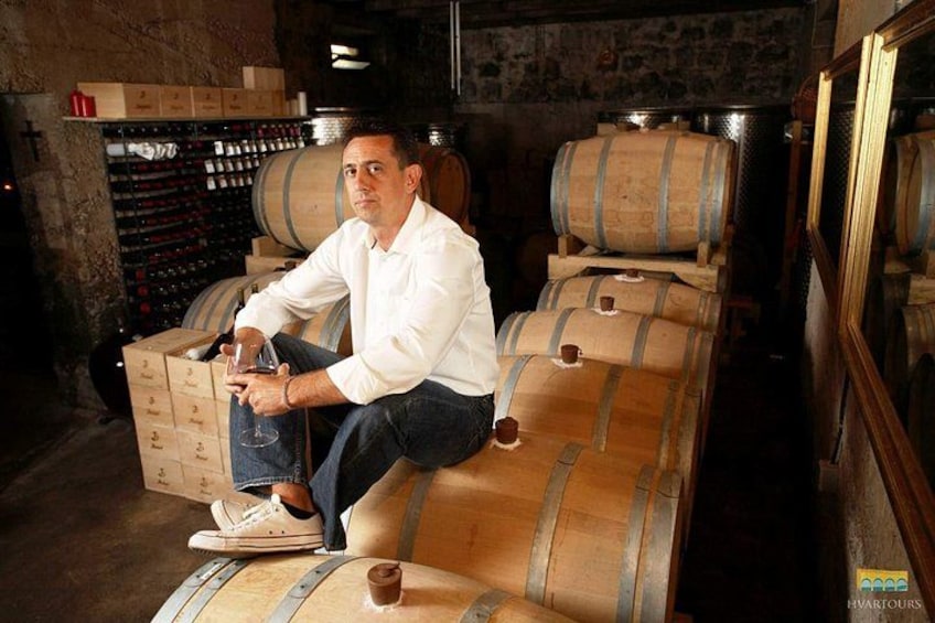 A person sitting on a barrel in a cellar with wine racks and barrels in the background. 3