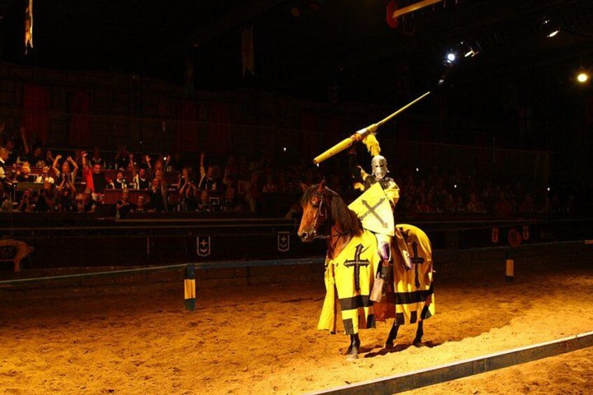 A knight in armor on horseback in a sandy arena with spectators. 3