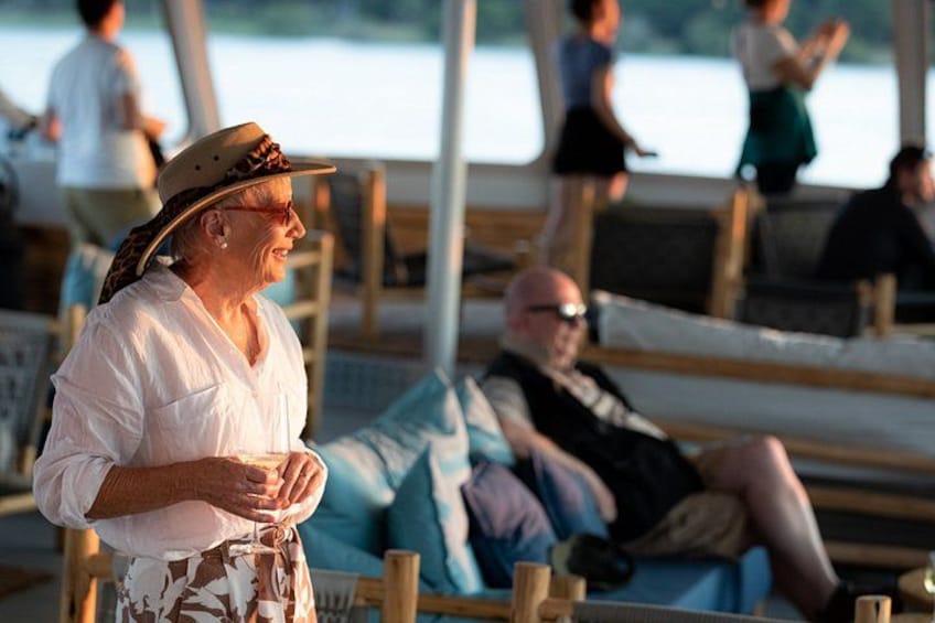 Une personne vêtue d’une chemise blanche et d’une jupe à motifs, debout sur une terrasse en bois, avec une vue sur l’eau et des personnes en arrière-plan. 4