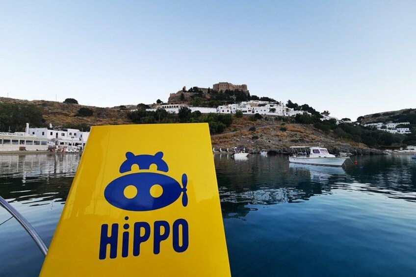 Een gele boot met een blauw logo en de tekst 'Hippo', liggend op het water, met witte gebouwen en een heuvel op de achtergrond. 5