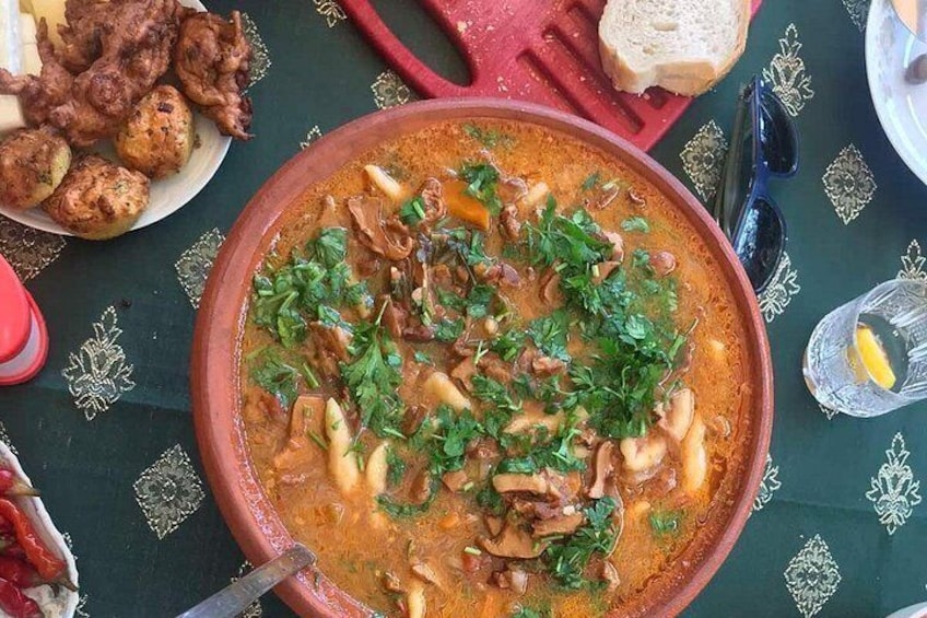 A large pot of stew with vegetables and herbs, a plate of bread, and a glass of water on a table with a patterned tablecloth. 5