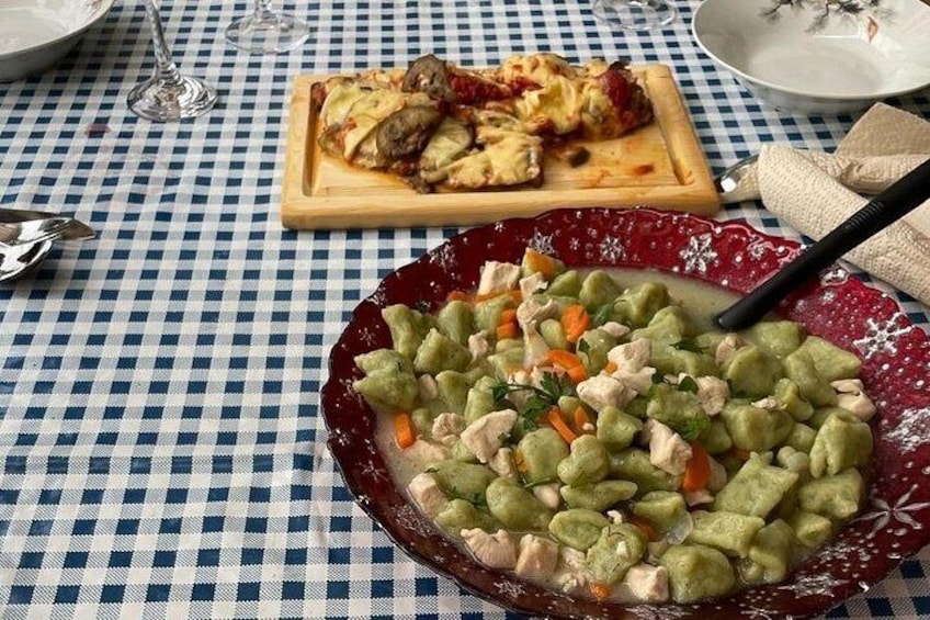 A table with a checkered tablecloth, a plate of green pasta with vegetables, a pizza on a wooden board, and a glass of wine. 3