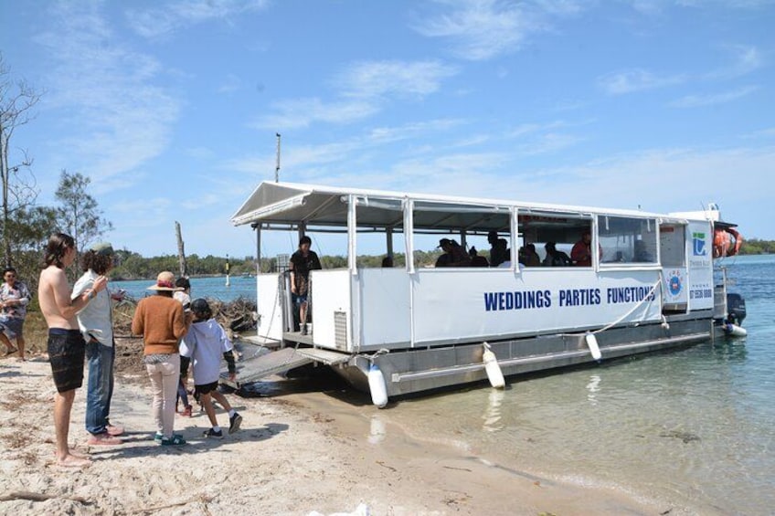 A boat with 'Weddings Parties Functions' written on it, docked at a sandy beach with people around. 3