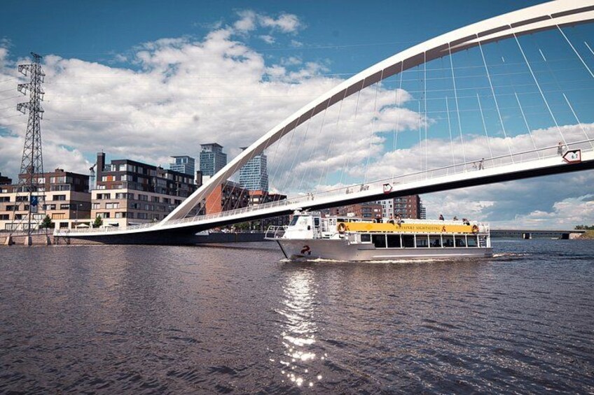Eine moderne Brücke mit einem Boot auf dem Wasser, im Hintergrund Stadtgebäude und ein klarer Himmel. 1