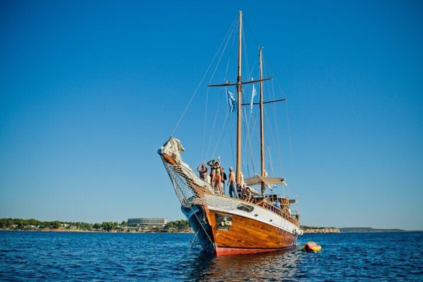 En træsejlskib med flere master på roligt vand, en klar himmel og en fjerntliggende kystlinje med bygninger. 3