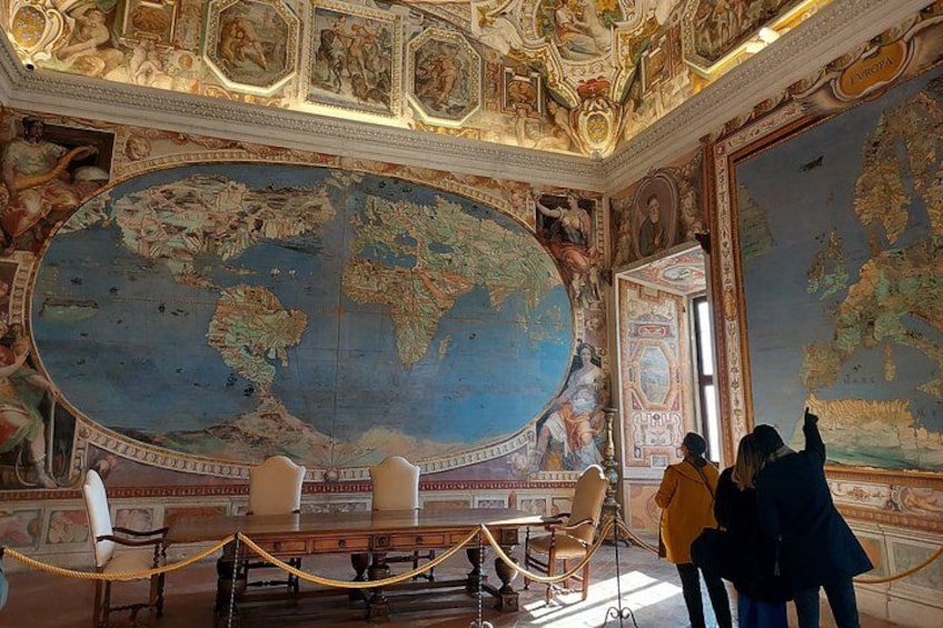 Een statige kamer met een grote wereldkaart op de muur, gedetailleerde plafondornamenten en een tafel met stoelen. 5