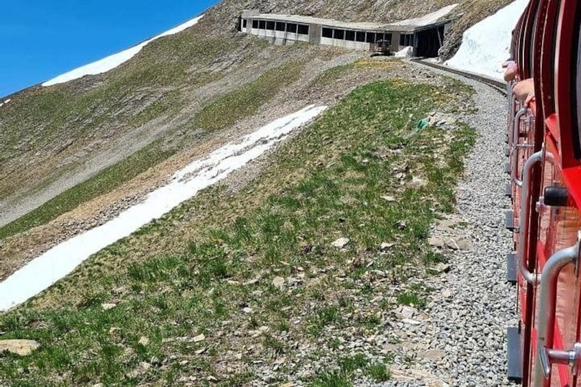 A train on a snowy mountain track, a building on the left, and a red train car on the right. 5