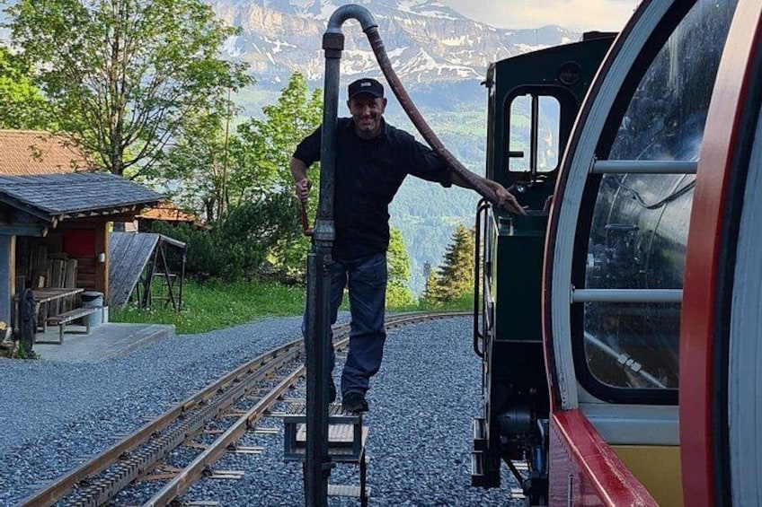 A man standing on a metal ladder next to a train car with a mountainous landscape in the background. 2