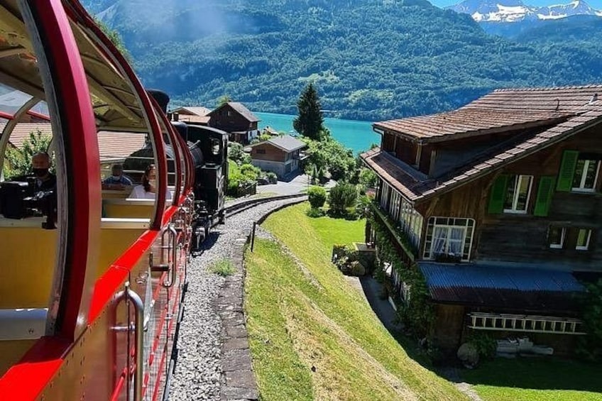 A red train on tracks, a wooden house, and a lake with mountains in the background. 1