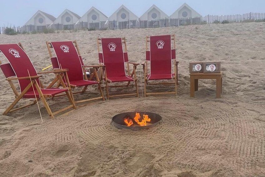 A beach with red lounge chairs, a small table, and a fire pit with flames. 2