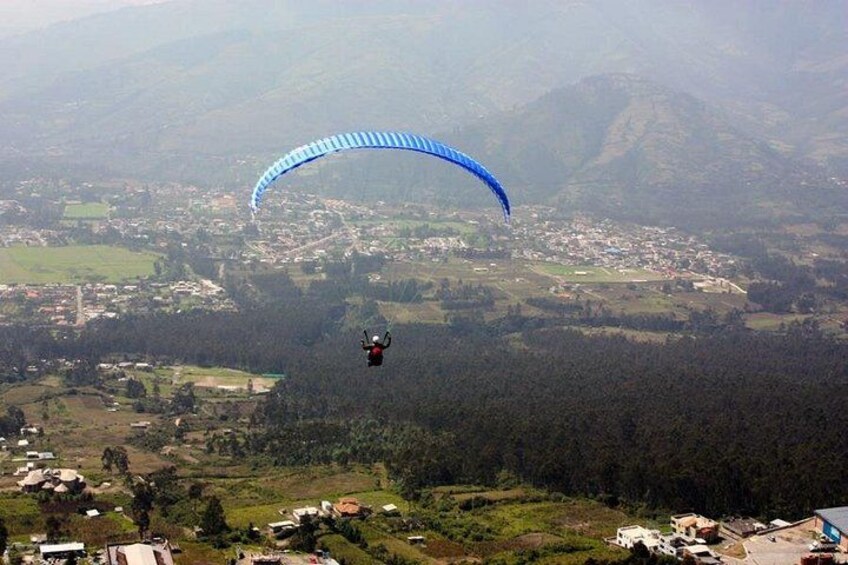 Een paraglider met een blauwe parachute vliegt boven een bergachtig landschap met een stad eronder. 4