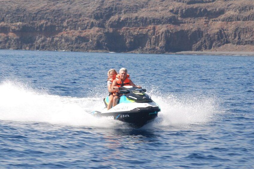 A jet ski with two people wearing life jackets on a body of water with a rocky shoreline in the background. 2