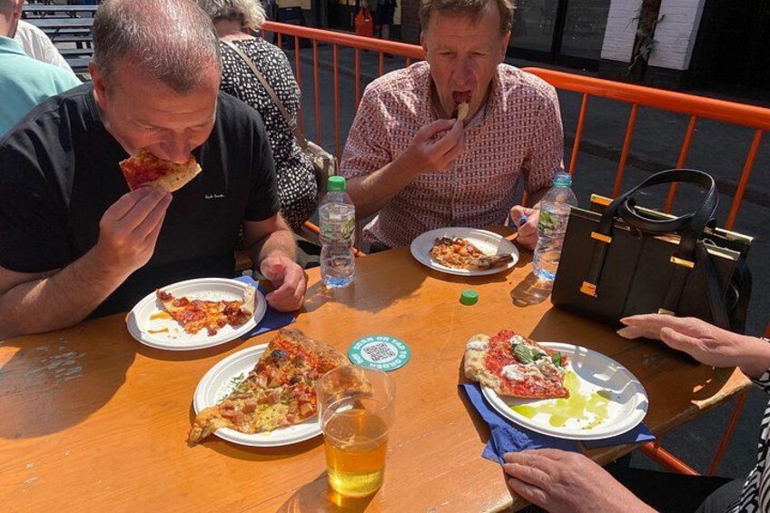 People eating pizza at an outdoor table with drinks and a handbag. 3