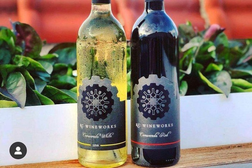 Two bottles of wine with labels, one labeled 'Crossroads White' and the other 'Crossroads Red,' both from KC Wineworks, placed on a wooden surface with greenery in the background. 2