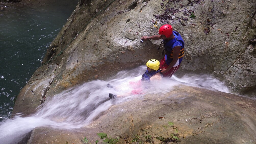 A person in a blue and red outfit and yellow helmet standing on a rock by a flowing river. 1