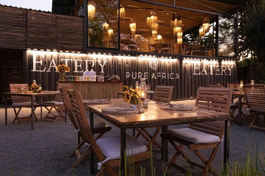 Un espacio de comedor al aire libre con mesas y sillas de madera, una barra con un letrero que dice 'EATERY PURE AFRICA', y luces colgantes. 2