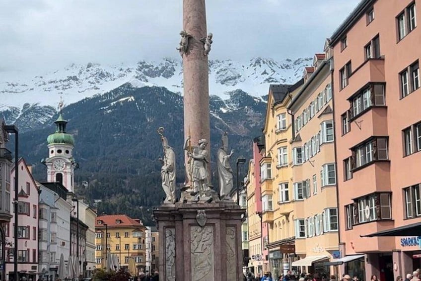 A town square with a tall monument, colorful buildings, and snow-capped mountains in the background. 1