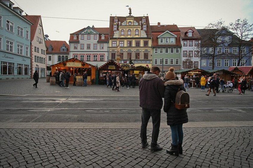 A cobblestone square with historic buildings, a market stall, and people walking. 2