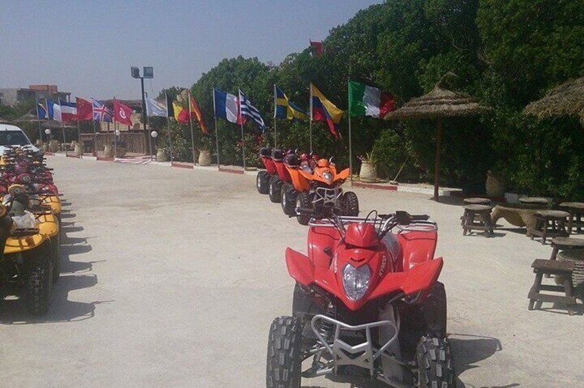A row of ATVs parked on a sandy area with flags and a thatched-roof structure in the background. 1