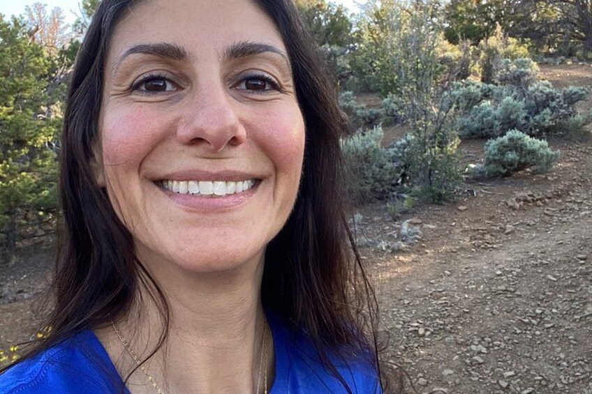 Una persona con cabello largo, usando una blusa azul, de pie al aire libre con un paisaje natural de fondo. 3