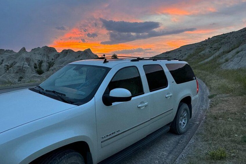 En hvid SUV parkeret på en grusvej med solnedgang i baggrunden. 5