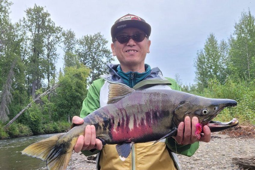 A person holding a large fish with pink markings, standing near a river with trees in the background. 4