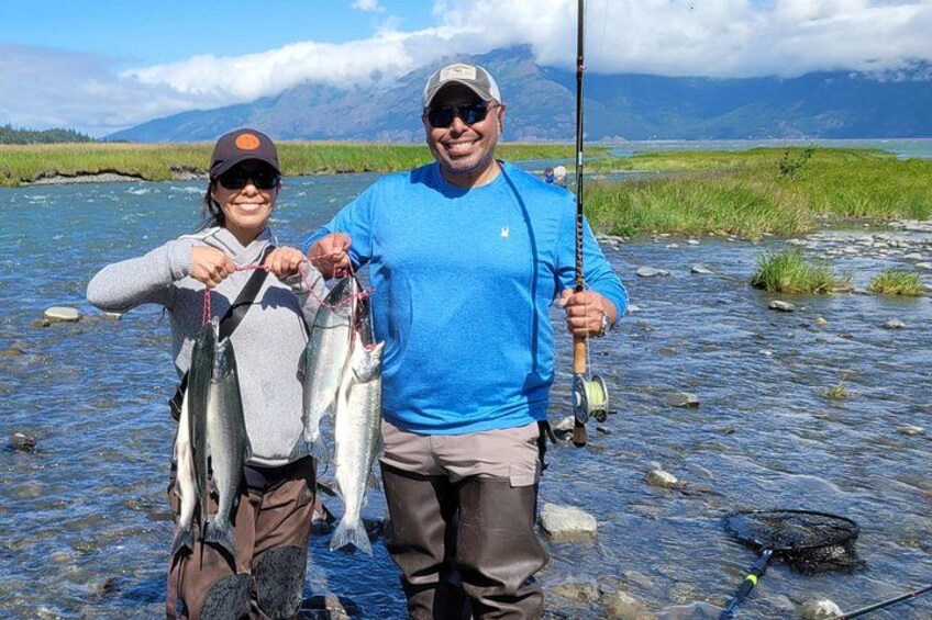 Two people holding fish, standing by a river with mountains in the background. 3