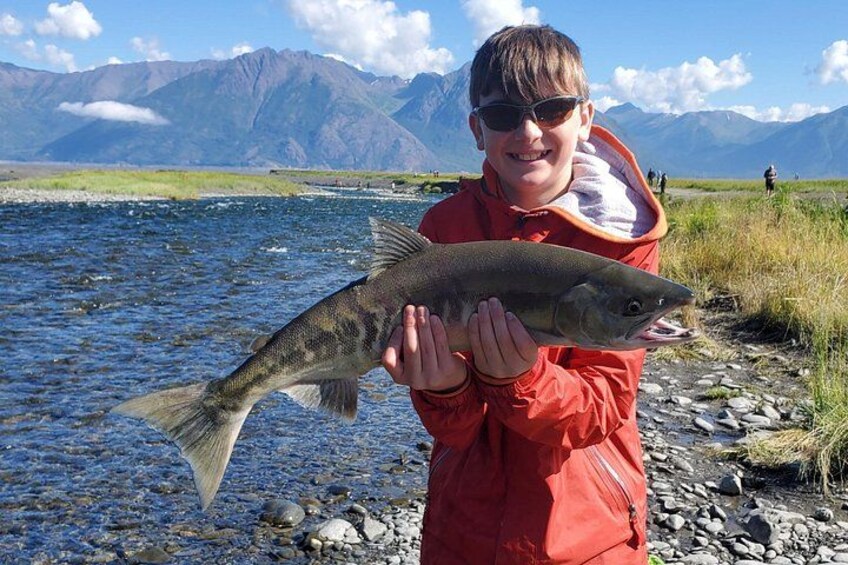A person holding a fish by a river with mountains in the background. 4